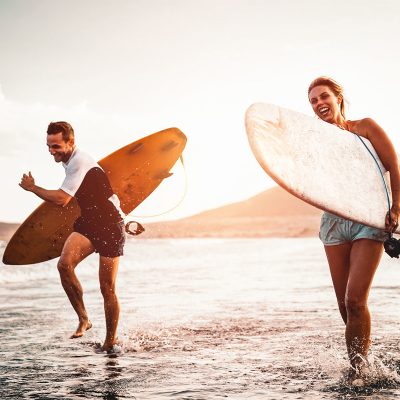 Happy surfer couple running with surfboards along the sea shore - Sporty people having fun going to surf together at sunset - Extreme surfing sport and youth relationship lifestyle concept