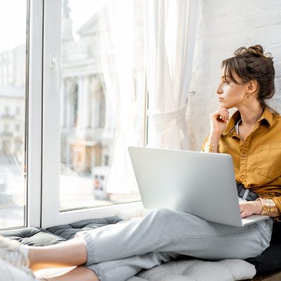 Young woman dressed casually working on laptop while sitting on the window sill at home. Work from home at cozy atmosphere concept